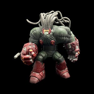 Op de afbeelding: Gedetailleerd actiefiguur van een gespierd, robotachtig personage. Het figuur is voornamelijk groen en rood, met een hoofd dat bestaat uit meerdere grijze buizen. Het figuur heeft grote, rode laarzen en een rode, mechanische arm.