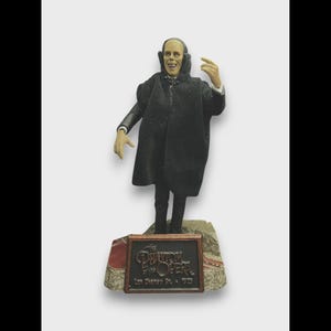 Peut inclure: Figurine du Fantôme de l'Opéra, vêtu d'un manteau et d'un costume noirs, avec un visage pâle et une expression sinistre. La figurine est posée sur un socle avec le titre et la date de la pièce.