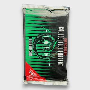 Könnte beinhalten: Versiegelte Sammelkartenpackung mit dem Wort "ALIENS" in Grün und einer stilisierten Alien-Kopf-Grafik. Die Packung ist schwarz mit grünen und roten Akzenten, und der Text "10 SAMMELKARTEN" ist sichtbar.