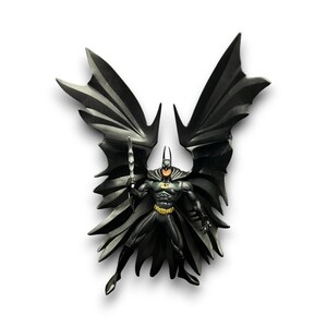 May include: A Batman figurine in a dynamic pose, with a black suit, yellow belt, and bat-shaped cape. The figure is holding a weapon and has a determined expression. The wings are spread wide, creating a dramatic silhouette.