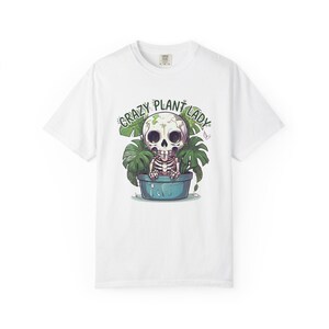May include: White t-shirt featuring a cartoon skeleton in a blue pot surrounded by green leaves. The text "CRAZY PLANT LADY" is arched above the skeleton. The design is centered on the shirt.