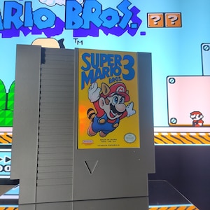 Puede incluir: Un cartucho de juego gris de Nintendo Entertainment System (NES) con una etiqueta amarilla. La etiqueta presenta una imagen de dibujos animados de Super Mario 3, con el texto "Super Mario 3 Bros." y "Version Española".
