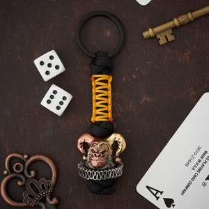 May include: A black and yellow paracord keychain with a bronze and gold jester monkey charm. The keychain is attached to a silver key ring.