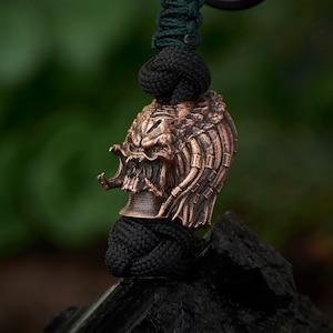 Puede incluir: Una cuenta de calavera Predator de color bronce, con detalles intrincados, cuelga de un cordón de paracord negro y verde. La cuenta está montada sobre una piedra negra, con un fondo verde borroso. La calavera presenta mandíbulas abiertas y rasgos faciales detallados.