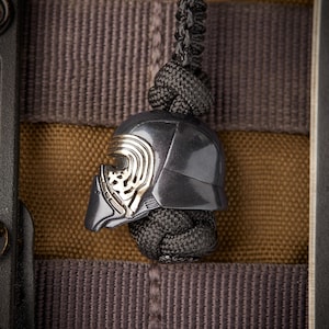 Könnte beinhalten: Ein schwarzer und silberner Kylo Ren Helm-Anhänger mit einem schwarzen Paracord-Lanyard. Der Anhänger ist an einem braunen und grauen Stoffhintergrund befestigt.