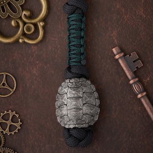 Op de afbeelding: Een zwarte en groene paracord sleutelhanger met een zilveren schildpadbedel. De schildpadbedel heeft een gedetailleerd schildontwerp.