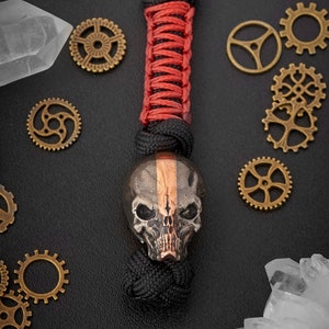 May include: A black and copper skull bead with a red and black paracord lanyard. The skull bead has a detailed design and a copper stripe down the center. The lanyard is made of braided paracord and has a knot at the end.