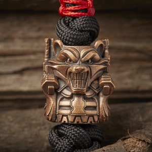 May include: A bronze-coloured metal charm featuring a detailed depiction of a raccoon with a fierce expression. The charm is attached to a black paracord bracelet with a red cord accent.