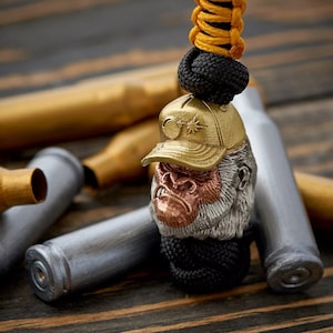 Pode incluir: Um amuleto de cabeça de gorila prateado e cobre com um boné de beisebol dourado no topo. O amuleto está preso a um cordão de paracord preto e amarelo.