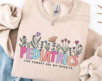 May include: Sand-colored sweatshirt with the word "PEDIATRICS" in colorful letters, above the phrase "tiny humans are my favorite." The design includes a floral border with various flowers. A silver necklace is visible.
