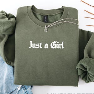 May include: An olive green crewneck sweatshirt featuring the white text Just a Girl printed on the chest. The garment has long sleeves with ribbed cuffs and a ribbed neckline. A silver-toned chain necklace with a circular pendant rests on the collar.