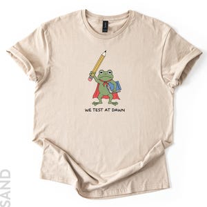 May include: Sand-colored t-shirt featuring a cartoon frog with a pencil, cape, and books. The frog is green with a red cape and holds a yellow pencil. The text "WE TEST AT DAWN" is printed below the frog.