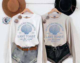 May include: Two long-sleeved sweatshirts with the text "Olivia's Bachelorette, Last Toast on the Coast, Miami, FL" and a seashell graphic. One is white, the other is beige. Both are paired with denim shorts and sunglasses.