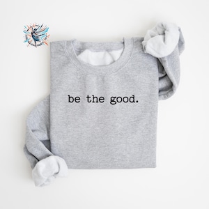May include: A light gray sweatshirt with the phrase "be the good" printed in black. The sweatshirt has a crew neck and long sleeves. The inside lining is white. A logo is visible in the upper left corner.