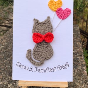 May include: A handmade greeting card with a crocheted grey cat wearing a red bow tie, holding yellow and pink heart-shaped balloons. The text "Have A Purrfect Day!" is printed below. The card is displayed on a small wooden easel.