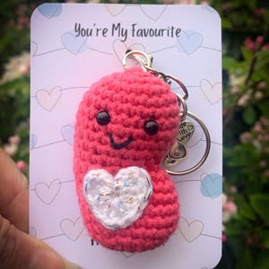 Puede incluir: Llavero de ganchillo en forma de frijol de color rosa con un corazón blanco y una cara sonriente. El llavero está unido a un llavero plateado con un dije que dice "Made with Love". El llavero se muestra en una tarjeta que dice "You're My Favourite".