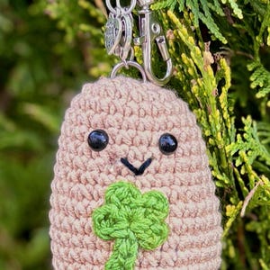 May include: A crocheted potato keyring with a smiling face, black bead eyes, and a green shamrock. The potato is tan and attached to a silver keyring with a lobster clasp. The background is green foliage.