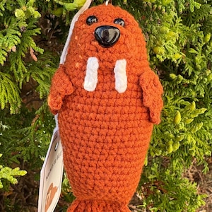 Puede incluir: Llavero de morsa naranja tejido a crochet con colmillos blancos y nariz negra. El llavero tiene brazos pequeños y aleta caudal. Una etiqueta con texto y un dije en forma de corazón que dice "Made with Love" están adjuntos.