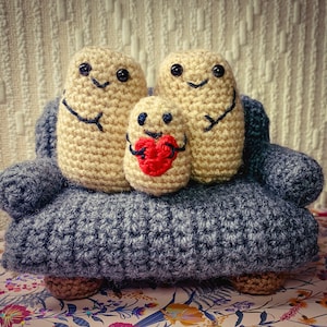May include: Three crocheted stuffed toys shaped like potatoes sitting on a grey crocheted sofa. The middle potato is holding a red heart.