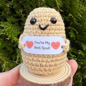 May include: A handmade crochet potato character with black button eyes and a stitched smile. It holds a sign that reads "You're My Best Spud" with heart accents. The figure is mounted on a small wooden base.