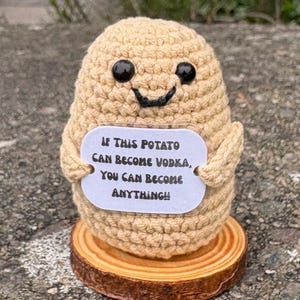 Puede incluir: Una figura de patata tejida a ganchillo en color beige con ojos de plástico negro y una sonrisa bordada, que sostiene un cartel blanco que dice: IF THIS POTATO CAN BECOME VODKA, YOU CAN BECOME ANYTHING!! La patata está sobre una base de madera.