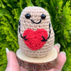 Pode incluir: Uma batata de crochê segurando um coração vermelho, em pé sobre uma fatia de madeira. A batata tem um rosto sorridente com olhos pretos.