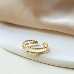 Peut inclure: Une bague dorée à double rangée avec un motif texturé et perlé. La bague est ouverte en haut et repose sur une surface circulaire blanche. L'arrière-plan présente un tissu beige doux.