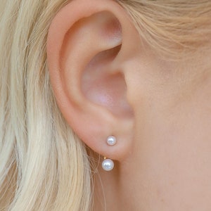 Pode incluir: Close-up de uma orelha com dois pequenos brincos de pérolas brancas. Os brincos apresentam uma pequena pérola branca na frente e outra atrás do lóbulo da orelha. Cabelo loiro. Design simples e elegante.
