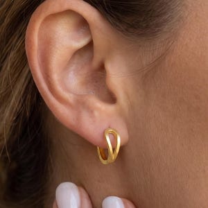 May include: A small gold hoop earring with a twisted, open-loop design. The earring features a smooth, polished finish and is worn in the earlobe. The hoop has a continuous, fluid shape that creates a subtle layered appearance on the ear.