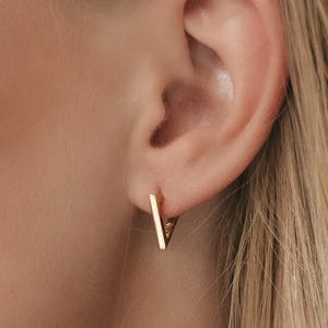 May include: Gold hoop earrings with a thin, straight bar design. The earrings are worn in a person's ear.