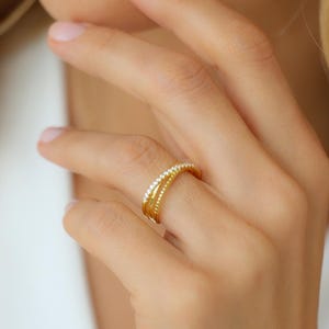 Könnte beinhalten: Nahaufnahme eines goldenen Rings mit einem Band aus kleinen, klaren Steinen. Der Ring wird an einem Finger getragen. Der Ring hat ein schlichtes, elegantes Design. Der Hintergrund ist verschwommen, mit Fokus auf dem Ring.