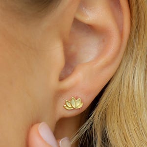 May include: A close-up of a person's ear with a small, gold-colored lotus flower stud earring. The earring is a detailed representation of the flower's petals. The image is well-lit, highlighting the earring's design and the ear's texture. The brand name "Piana Me" is visible.