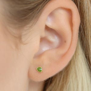 May include: A close-up of a person's ear with a small green gemstone earring. The earring is a simple stud style with a gold setting.