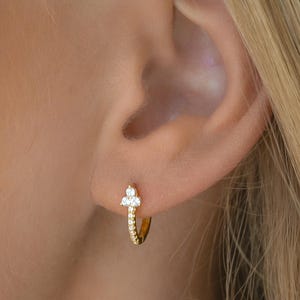 May include: Gold hoop earrings with a small cluster of clear cubic zirconia stones. The earrings are worn in a person's ear.