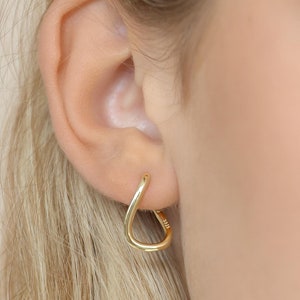 Puede incluir: Un par de pendientes de aro dorados con forma de lágrima. Los pendientes se llevan en la oreja de una persona con el pelo rubio.