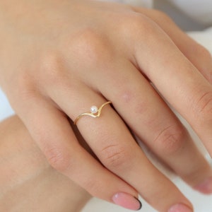 Peut inclure: Une bague en or avec un petit accent de perle. La bague est en forme de V et est portée sur un doigt.