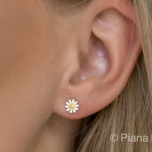 May include: A pair of silver daisy stud earrings with a yellow center. The earrings are shown in a close-up image of a person's ear.