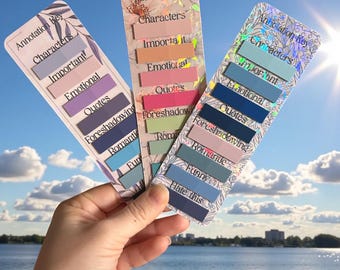 May include: Three colorful annotation key bookmarks are held up against a blue sky. Each bookmark has a different design with text labels like "Characters," "Important," and "Quotes." The bookmarks are designed for annotating books.