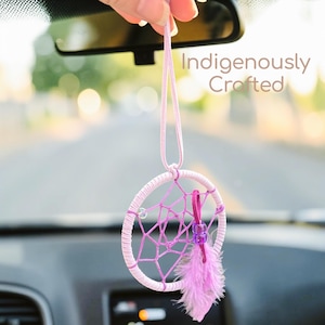 May include: A handcrafted dreamcatcher in shades of pink and purple, hanging from a string. The dreamcatcher features a circular frame with a webbed center, a purple feather, and the text "Indigenously Crafted".