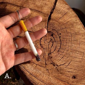 May include: A beaded cigarette-shaped keychain. The keychain features a yellow, white, and black beaded design, with a silver-colored metal keyring. The letter "A" is visible in the lower left corner.