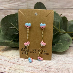 May include: A pair of dangle earrings with three pink and blue heart charms on each earring. The hearts are layered on a gold chain. The earrings are displayed on a brown card with the text "TURTLE CAY CRAFT STUDIO".