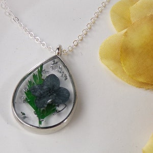 Puede incluir: Un collar de cadena de plata con un colgante en forma de lágrima. El colgante contiene una flor azul oscuro prensada con hojas verdes y purpurina plateada.