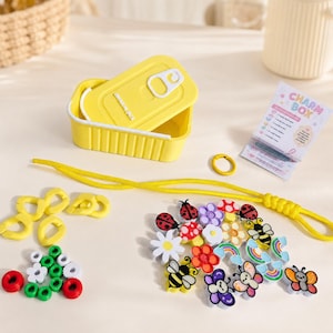 Op de afbeelding: Een gele rechthoekige container met het label CHARM BOX en een deksel, een geel koord, een gele ring en een verzameling kleine kleurrijke bedels in de vorm van lieveheersbeestjes, bijen, bloemen, paddenstoelen en regenbogen, plus kleine groene, witte en rode ringen.