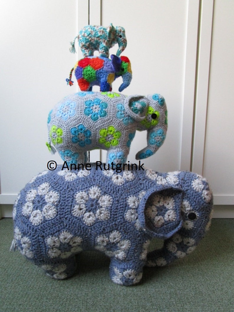 Op de afbeelding: Een stapel van drie gehaakte olifantenbeeldjes. De grootste is blauw met witte bloempatronen. De middelste olifant is veelkleurig en de kleinste is lichtblauw met een bloemmotief. Handgemaakt en decoratief.