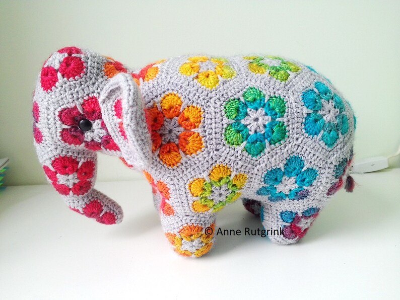 Op de afbeelding: Een handgemaakte gehaakte olifant, versierd met kleurrijke bloempatronen in rood, oranje, geel, groen en blauw. De olifant heeft een grijze basis en een knoop als oog.