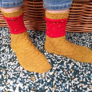Puede incluir: Un par de calcetines de punto en amarillo y rojo. Los calcetines tienen un patrón de punto de cable y están decorados con pequeñas cuentas negras.