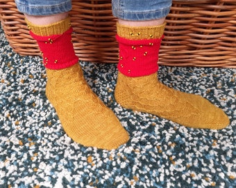 Op de afbeelding: Een paar gebreide sokken in geel en rood. De sokken hebben een kabelbreipatroon en zijn versierd met kleine zwarte kralen.