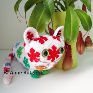 Peut inclure: Un chat en crochet blanc avec des fleurs rouges, vertes, bleues et roses sur son corps. Le chat a des yeux verts et un visage blanc. Le chat est assis sur une surface blanche.