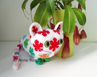 Op de afbeelding: Een witte gehaakte kat met rode, groene, blauwe en roze bloemen op zijn lichaam. De kat heeft groene ogen en een wit gezicht. De kat zit op een wit oppervlak.