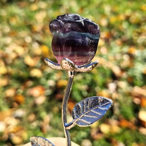 May include: A handcrafted, gemstone rose sculpture. The rose is carved from a purple and green fluorite crystal, mounted on a silver-colored metal stem with detailed leaves. The sculpture is held against a blurred background of green and yellow.